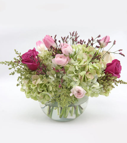 Blooms of Hope bouquet features lavender roses, green hydrangea, pink ranunculus, pink tulips, limonium and delicate hints of foliage. It is a charming fishbowl vase arrangement.